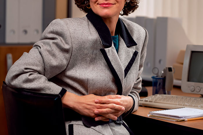 Woman wearing vintage blazer seated at office desk with computer and notepad, evoking stirrup pants fashion trends.