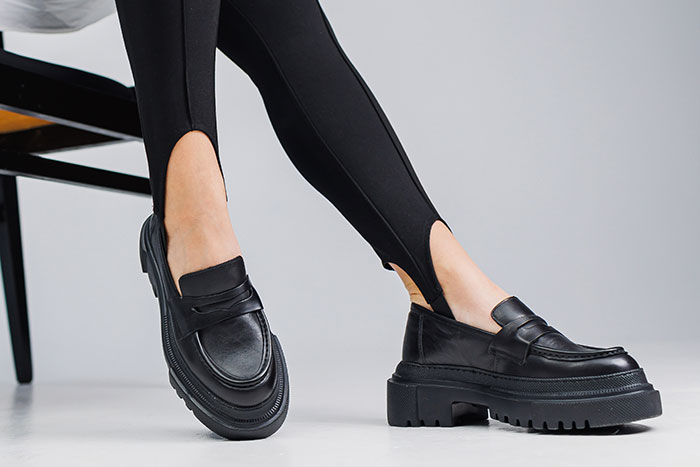Person wearing black stirrup pants and chunky black loafers seated on a chair against a plain background.
