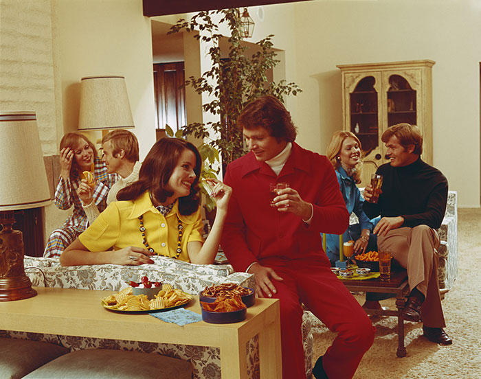 Group of young people socializing in vintage clothes, some wearing stirrup pants at a casual 1970s indoor gathering.