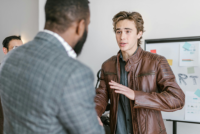 Young worker in a brown leather jacket discussing the one minute late rule with his boss in an office setting.
