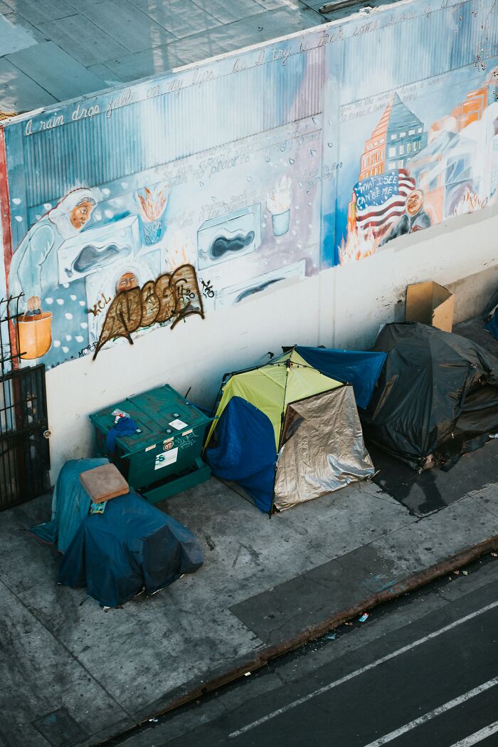 Outdoor urban scene showing tents set up on a sidewalk under a mural capturing out-of-touch people shamed online.
