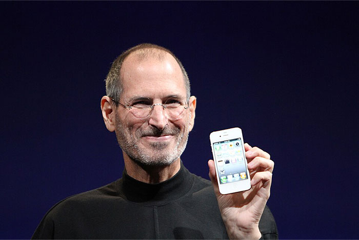 Steve Jobs smiling in a black turtleneck, holding up a white smartphone during a product presentation.