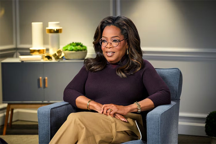Oprah Winfrey seated in a chair wearing glasses and a dark top, in a well-lit modern interview setting.