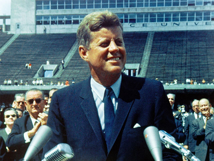 John F. Kennedy speaking at a crowded stadium event, symbolizing public figures praised after passing.