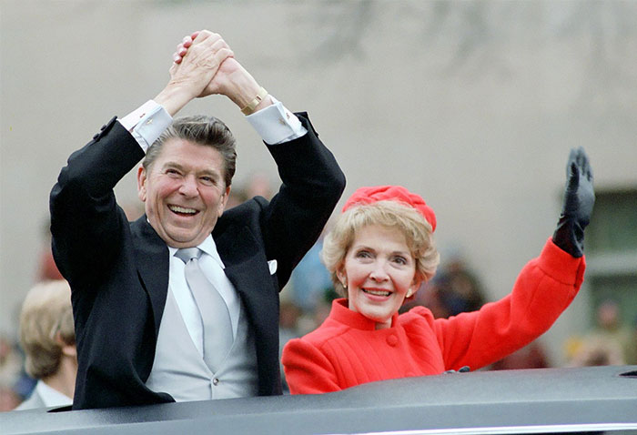 Former President Ronald Reagan and Nancy Reagan waving to a crowd during a public event, smiling and dressed formally.