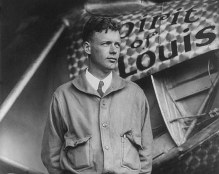 Black and white photo of a young man standing in front of a vintage airplane with Spirit of St. Louis written on it.