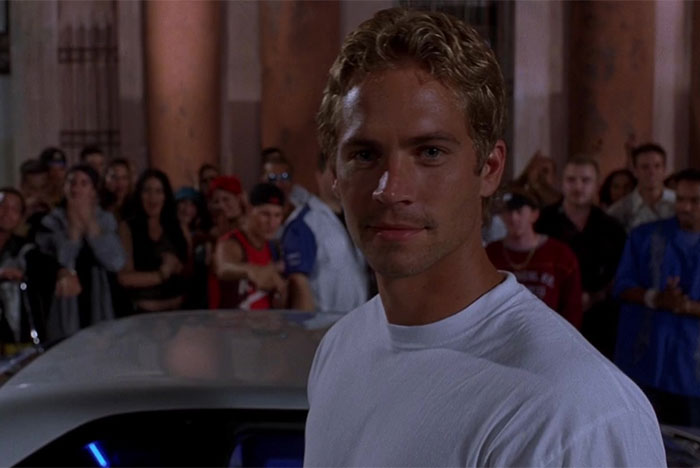 Young man in a white shirt standing in front of a car with a crowd behind, reflecting on Steve Jobs and celebrity legacy.