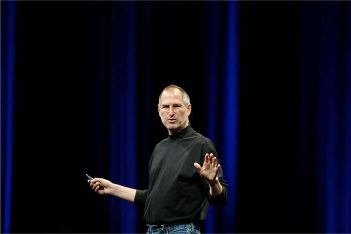 Steve Jobs speaking on stage wearing a black turtleneck and jeans during a presentation with blue curtains backdrop
