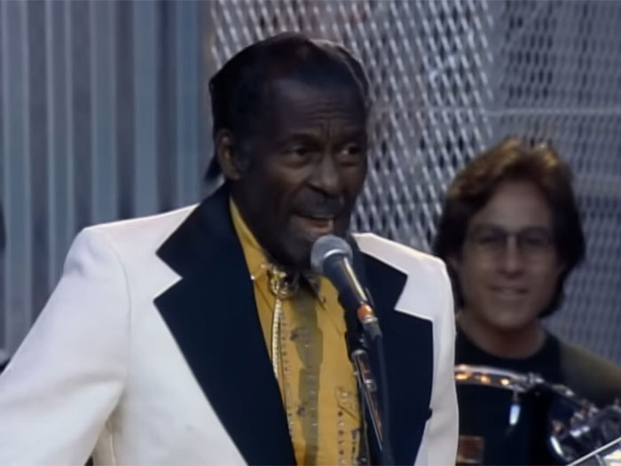Man in a white suit with black lapels singing into a microphone, with a drummer in the background, related to Steve Jobs topic.