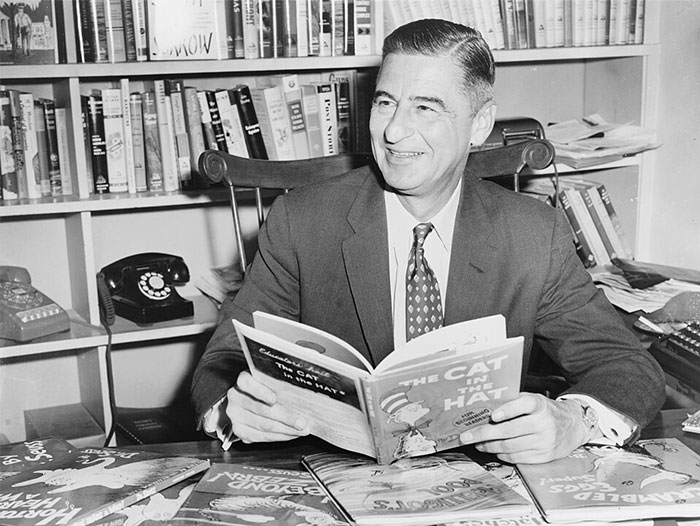 Man in a suit reading The Cat in the Hat book, surrounded by children’s books in a room with bookshelves.