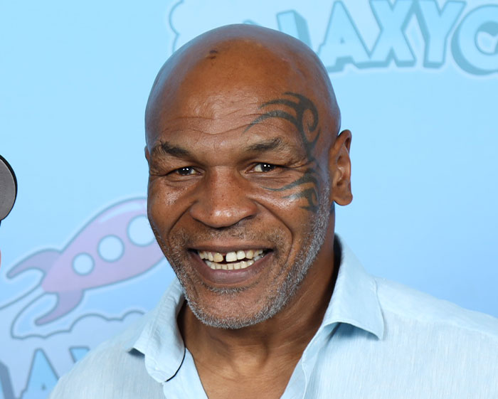 Smiling man with facial tattoo wearing a light blue shirt at an event with a blue background featuring logos.