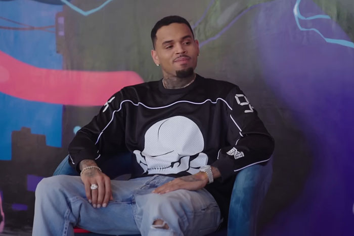 Man with tattoos wearing a black shirt with a skull design, sitting in a blue chair against colorful wall art.