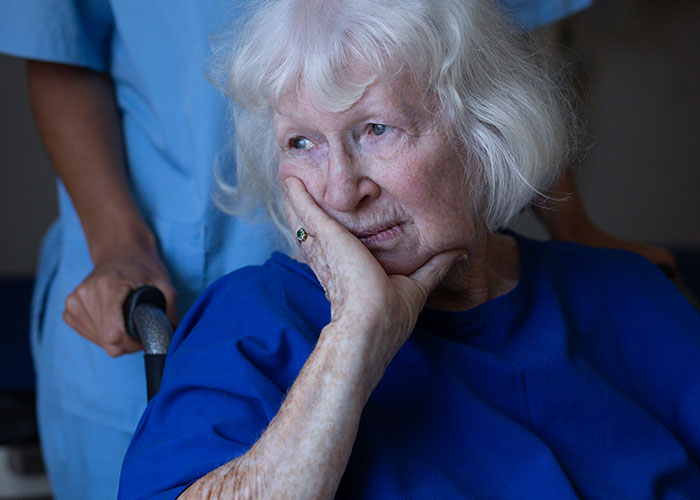 Elderly woman in a wheelchair looking contemplative with a nurse behind her, illustrating doctors and nurses final words.