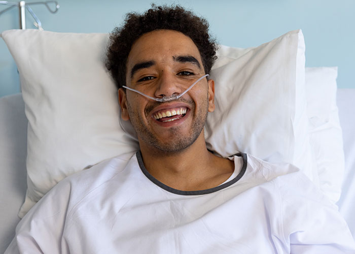 Young male patient in hospital bed with nasal oxygen, smiling as doctors and nurses hear his final words.
