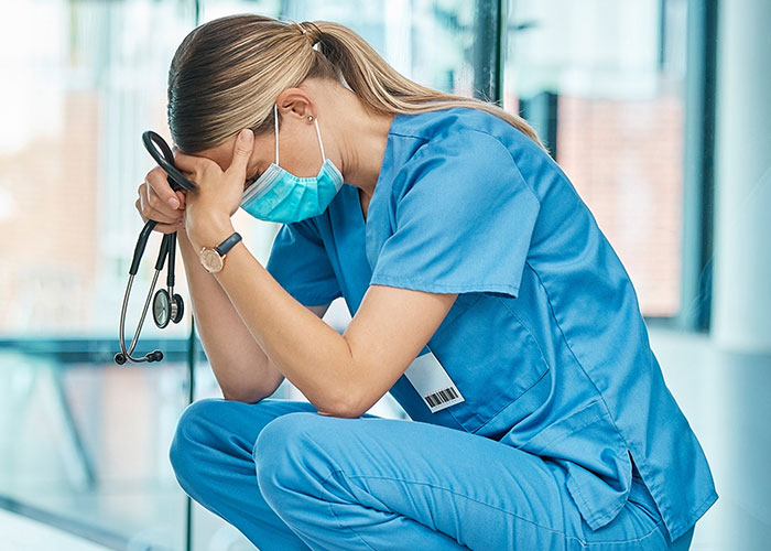 Female nurse in blue scrubs and mask sitting with head in hands, overwhelmed by chilling final words from dying patients.