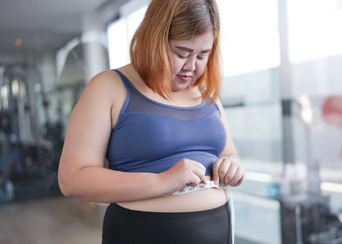 Woman in workout clothes measuring waist with tape, emphasizing patient’s lie impact on health and medical risks.