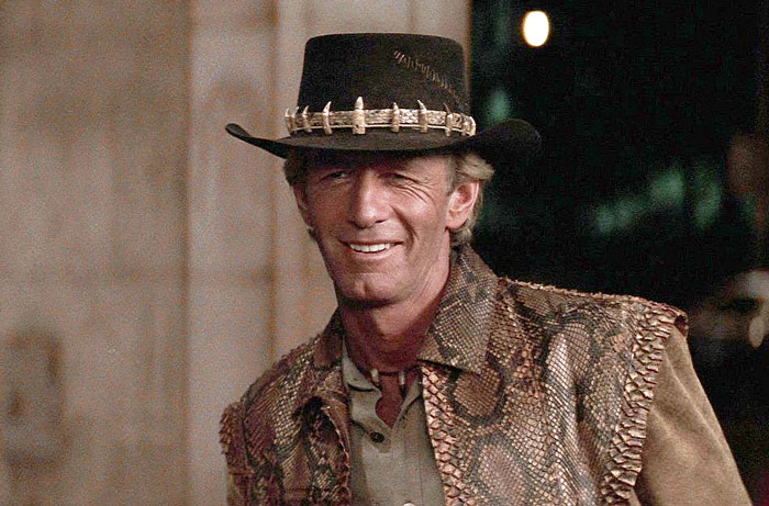 Paul Hogan as Crocodile Dundee wearing a crocodile tooth hat and snakeskin vest, smiling in a movie scene.