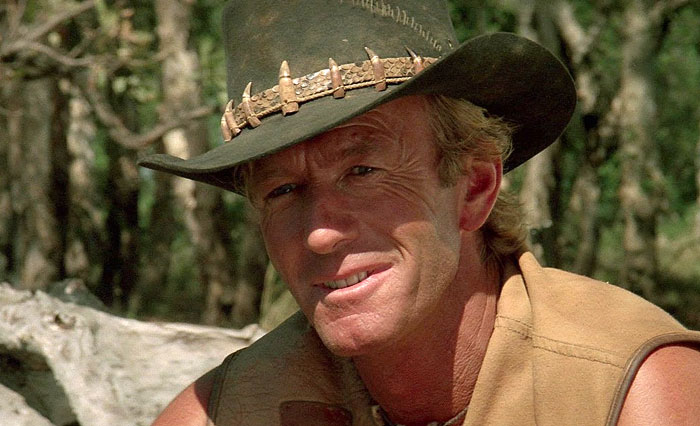 Paul Hogan as Crocodile Dundee wearing a hat with teeth, smiling in an outdoor forest setting.