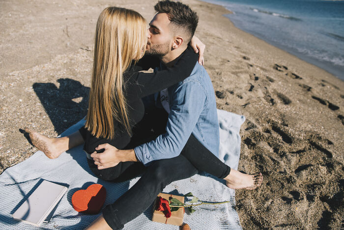 Couple sharing a romantic beach kiss sitting on a blanket with gifts and a rose, illustrating romanticized experiences.