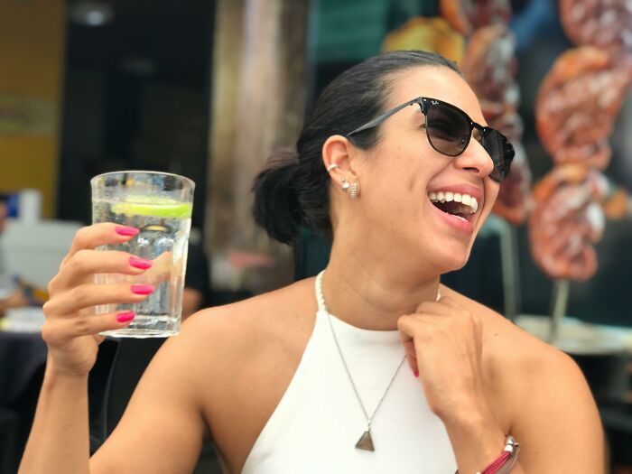 Woman laughing outdoors wearing sunglasses and holding a glass of water, illustrating romanticize experiences concept.