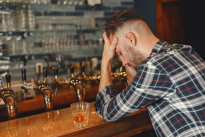 Man with head in hands sitting at bar counter with alcohol glass, illustrating folks only romanticize things they have not experienced.