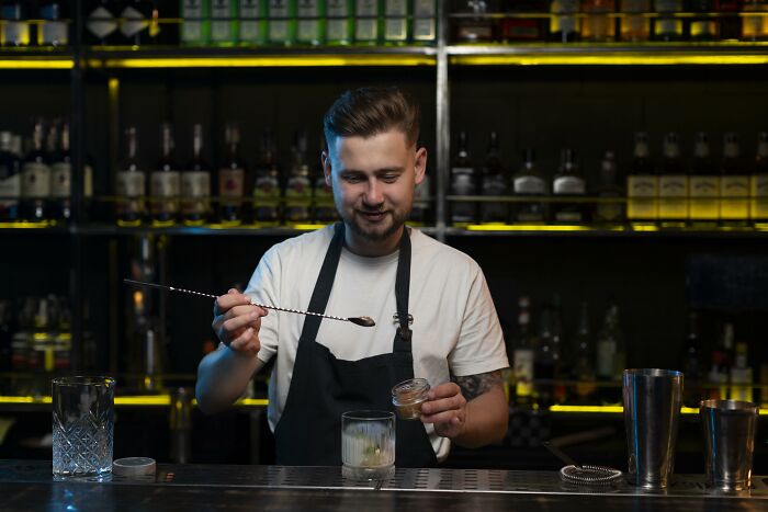 Bartender in a black apron preparing a cocktail behind the bar, representing things folks only romanticize.