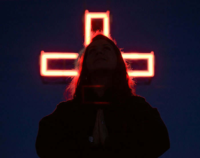Silhouetted person praying in front of a glowing red cross, reflecting the experiences of former cult members in dark setting.