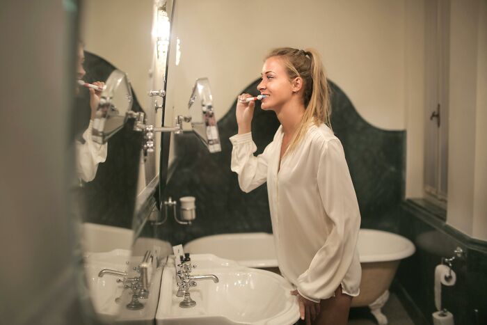 Young woman brushing teeth in bathroom mirror, highlighting daily habits related to tinnitus health awareness.