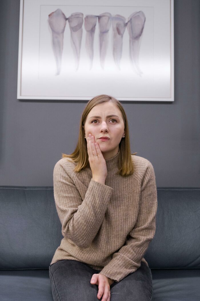 Woman in beige sweater sitting on couch, holding her cheek with a worried expression, indicating discomfort and tinnitus pain.