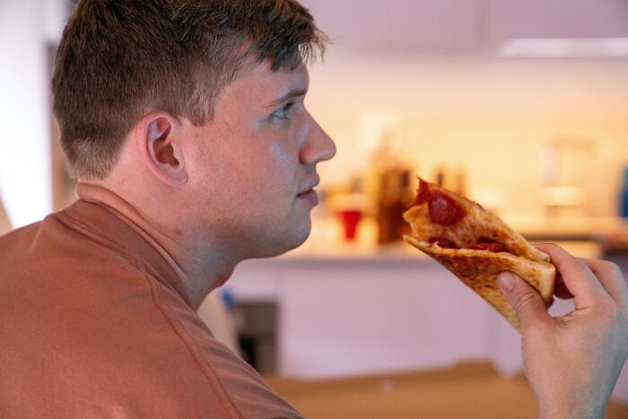 Young man holding a slice of pepperoni pizza, indoors, illustrating lifestyle choices linked to tinnitus on a daily basis.