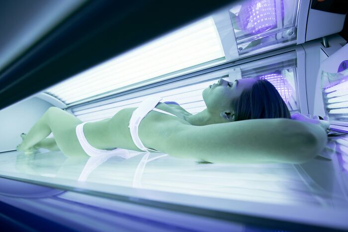 Young woman in white bikini lying inside a tanning bed under bright UV lights, relaxing with eyes closed.