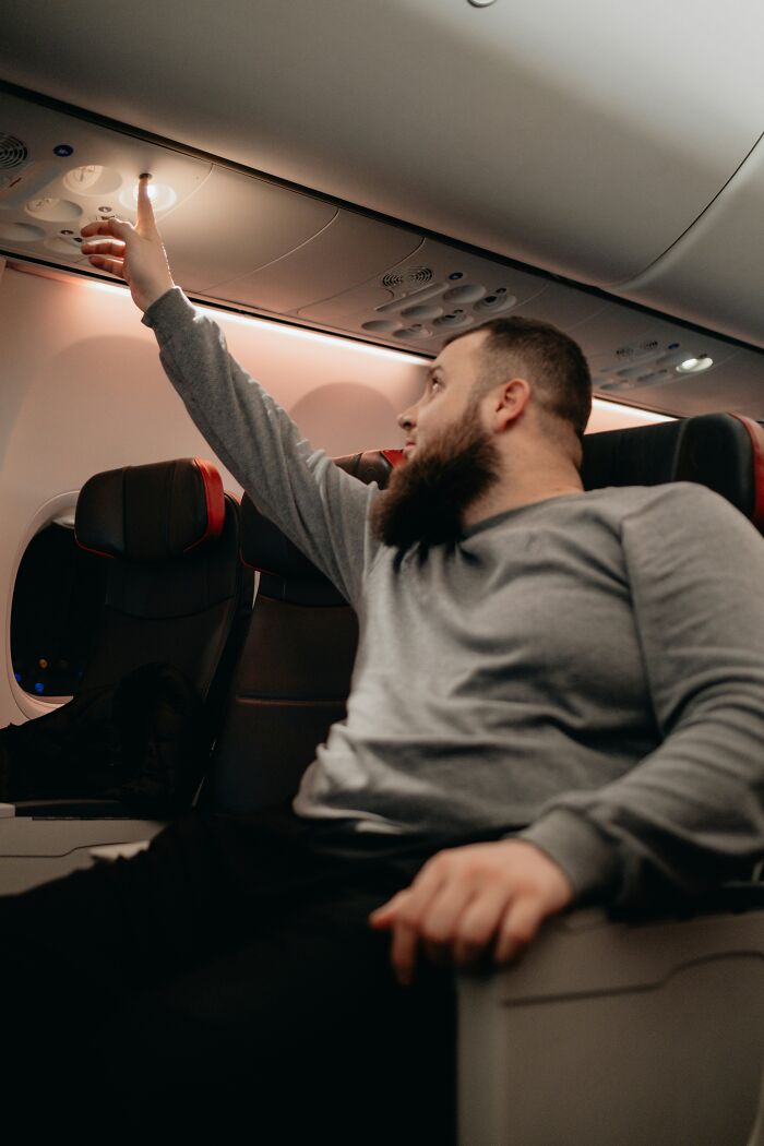 Man with beard reaching for airplane overhead controls while seated, illustrating microfeminisms in everyday settings.