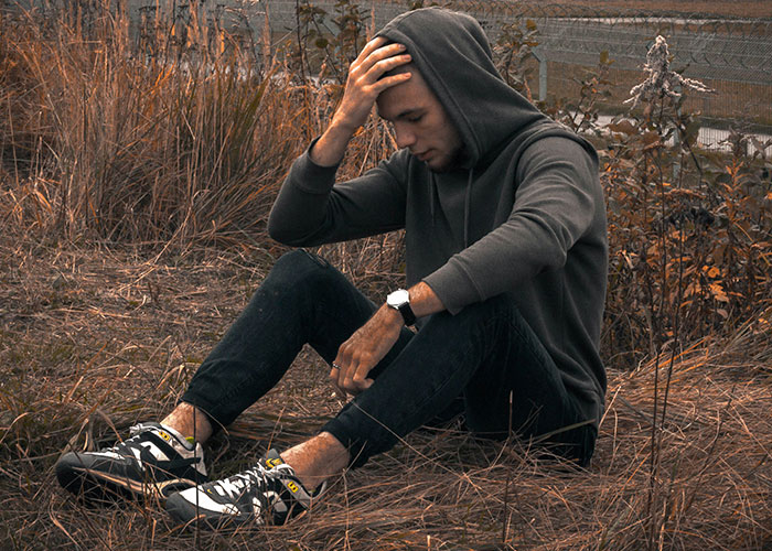 Young man in a hoodie sitting on dry grass, holding his head, representing emotions after exposing cheating partners.