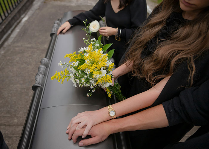 Two women dressed in black holding hands and flowers, standing beside a gray coffin at a funeral.