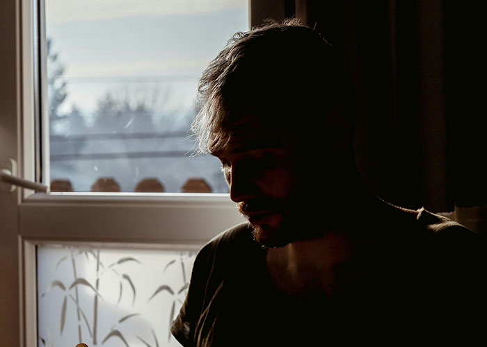 Silhouetted man sitting near window in low light, reflecting on trust and cheating partners in a dim room.
