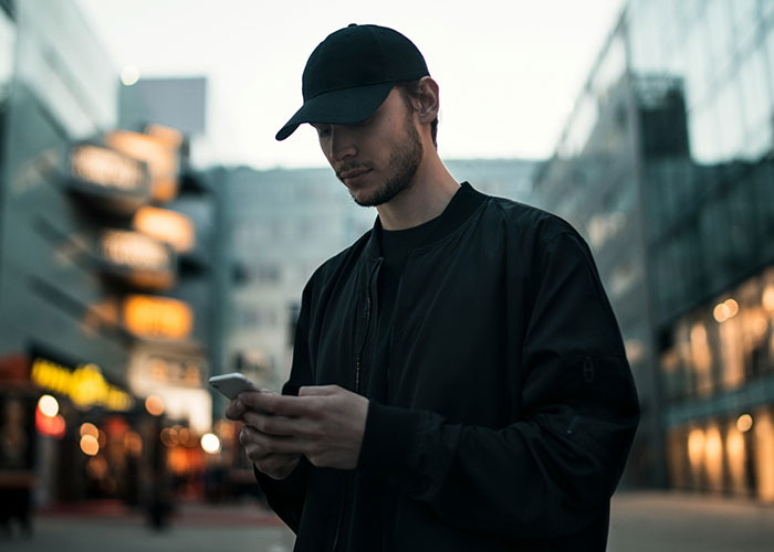 Young man in black jacket and cap texting on phone outdoors, capturing the moment of exposing cheating partners.