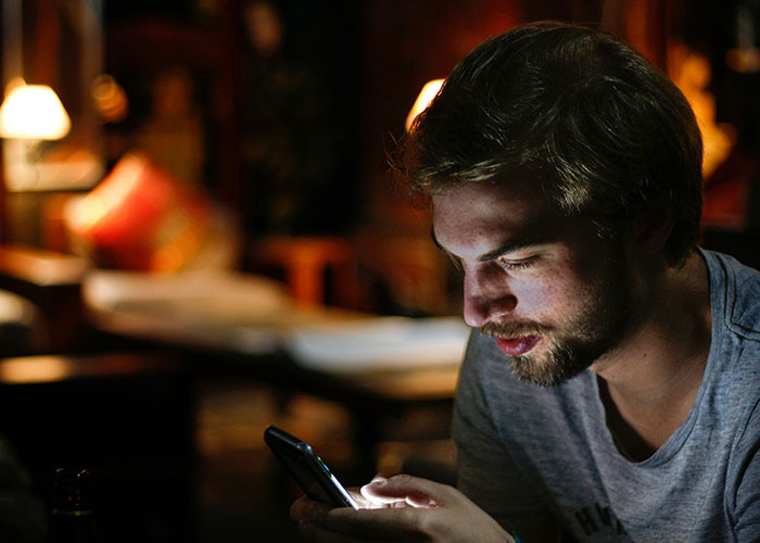 Young man in a dimly lit room looking at his phone, symbolizing how people reveal cheating partners online.