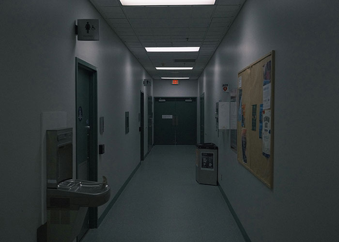 Dimly lit hospital corridor with water fountain, bulletin board, and restroom signs along the walls at night.