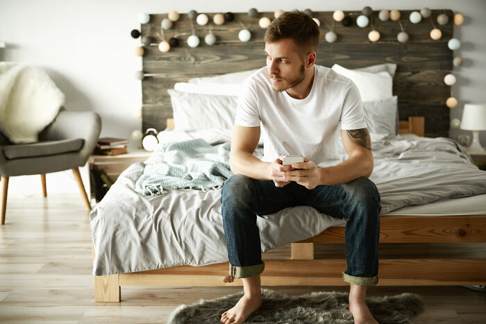 Young man sitting on bed in modern bedroom, holding phone, reflecting on spooky tales of resurrection and time travel.
