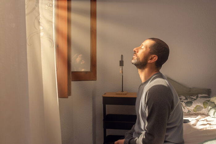 Man sitting on bed with eyes closed in warm sunlight, evoking a feeling of time travel and resurrection.