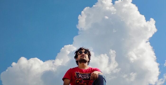 Man in sunglasses and red shirt against blue sky with large clouds, evoking spooky tales of resurrection and time travel.