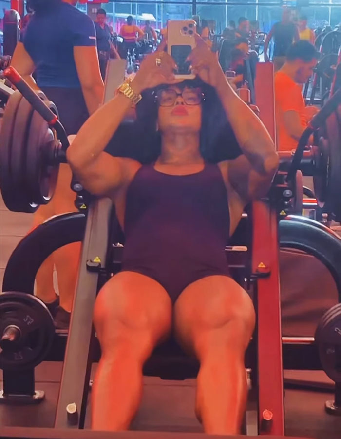 Woman using gym leg press machine, strong build, taking a selfie, scene focused on gym bathroom confrontation keywords.