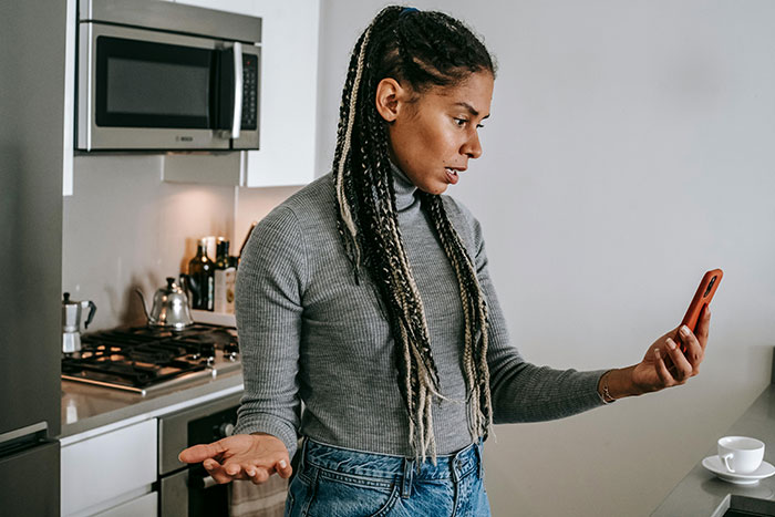 Woman in kitchen looking frustrated at phone, reacting to husband guarding box of olives like precious gold.