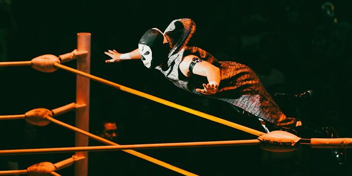 Wrestler wearing mask and costume performing shockingly real aerial move over ring ropes during intense wrestling event.