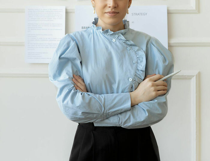 Teacher in ruffled light blue blouse holding a pen with arms crossed, demonstrating unhinged but effective teaching methods.