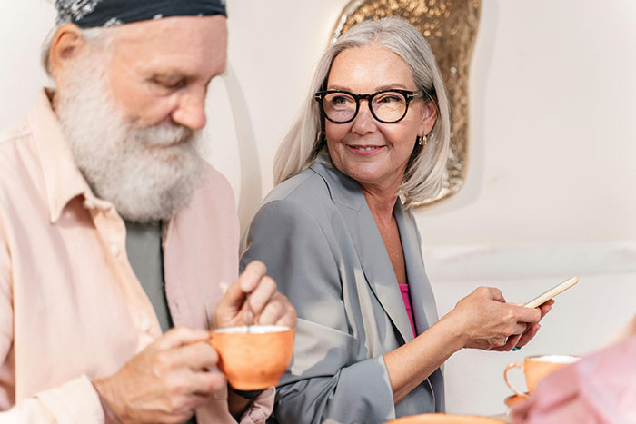 Older woman smiling while holding phone, sitting with older man drinking coffee, highlighting dog mom dilemma. Older woman smiling while holding phone, sitting with older man drinking coffee, highlighting dog mom dilemma.