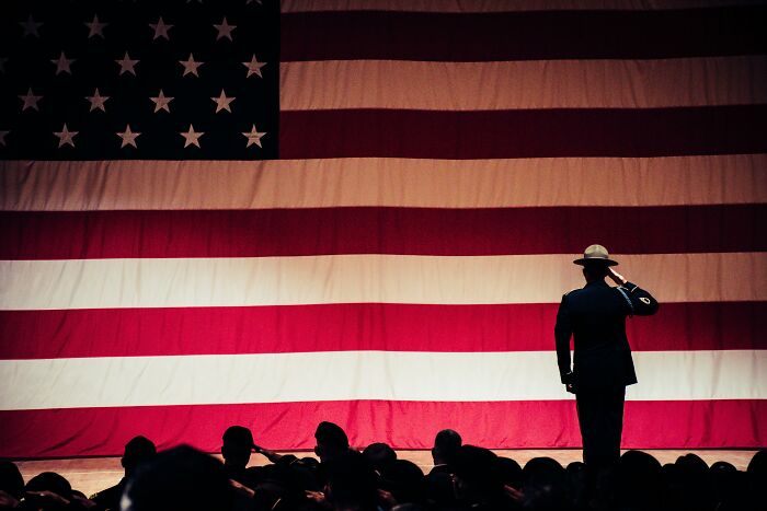 A WWI recruit saluting a large American flag with a crowd silhouetted in front during a military test or ceremony.