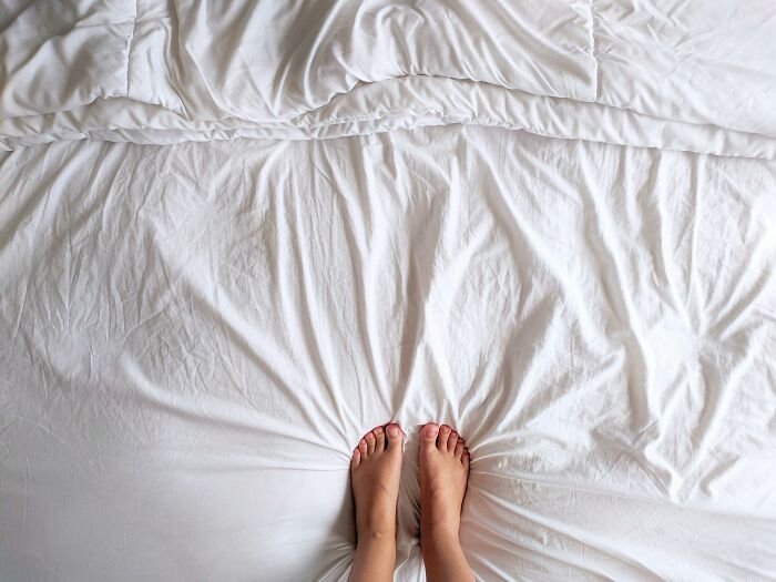 Bare feet resting on a soft, wrinkled white bed sheet, illustrating simple low-key pleasures of happiness.