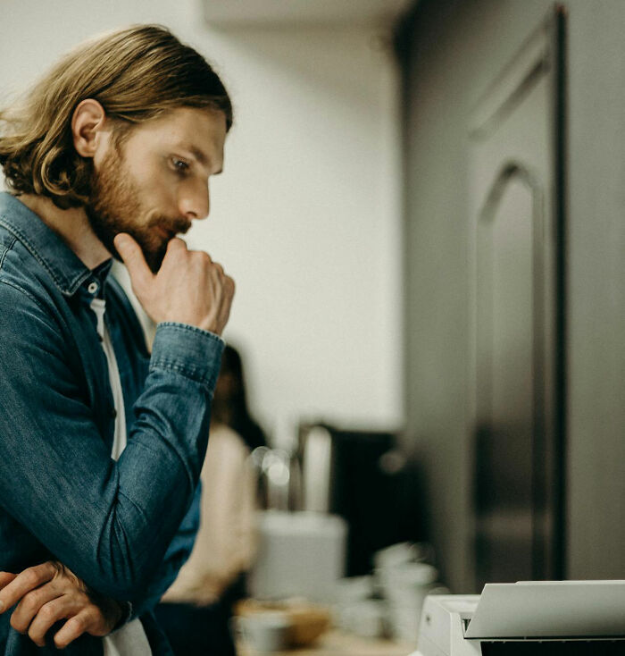 Man with beard and long hair looking thoughtfully at a printer, expressing concern about feeling instantly unsafe with a man.