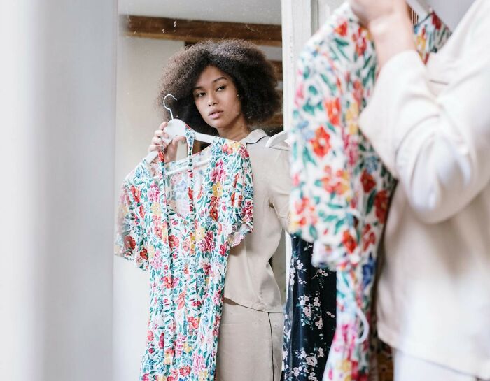 Young woman holding a floral dress in front of a mirror, illustrating a moment of excessive laziness.